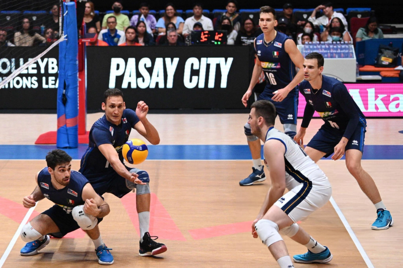 Volleyball players in blue uniforms are in action during a match. One player is diving to dig the ball, with teammates positioned nearby. A scoreboard and audience are visible in the background.