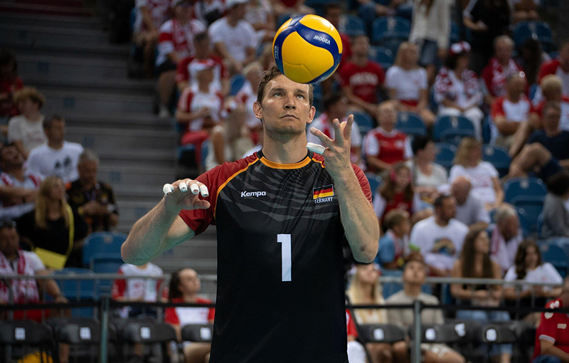 A volleyball player wearing a black and red jersey with the number 1, focusing on a volleyball he is tossing in the air. The audience is visible in the background.