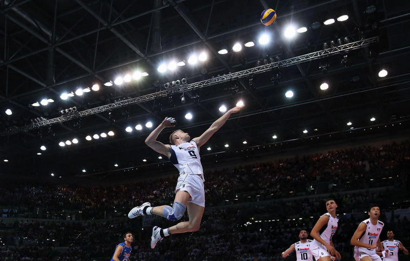 A volleyball player in a white uniform mid-air, preparing to spike the ball, with teammates and a crowded arena in the background.
