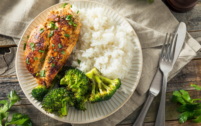Grilled chicken breast with herbs, white rice, and broccoli on a plate with a fork and knife set beside it.