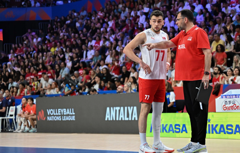 A volleyball coach gives instructions to a player wearing the number 77 jersey during a game at the Volleyball Nations League. The court is surrounded by a large crowd of spectators.