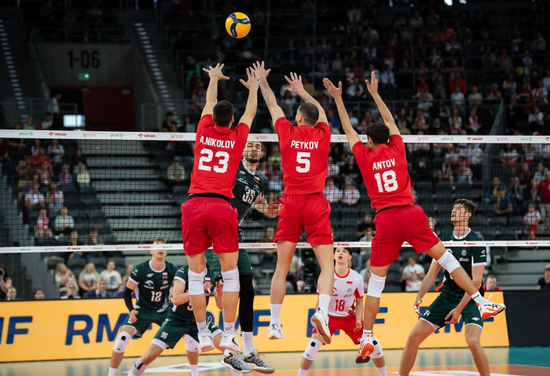 Three volleyball players in red jerseys jump to block a spiking player in green, with a filled stadium in the background.