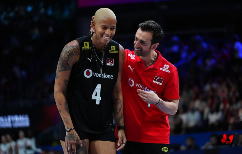 A volleyball player wearing a black jersey with the number 4 is smiling while interacting with a person in a red Turkish team jacket. They appear to be on an indoor court with an audience in the background.