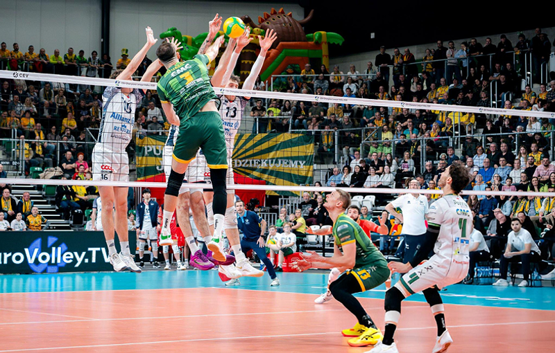 Volleyball players in mid-air at the net, with one team in green uniforms and the other in white. The crowd watches intensely from the stands.