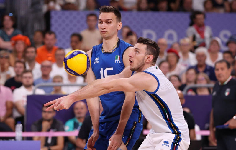 Two volleyball players in blue and white uniforms from Italy are focusing on the ball during a game, with a blurred crowd in the background.