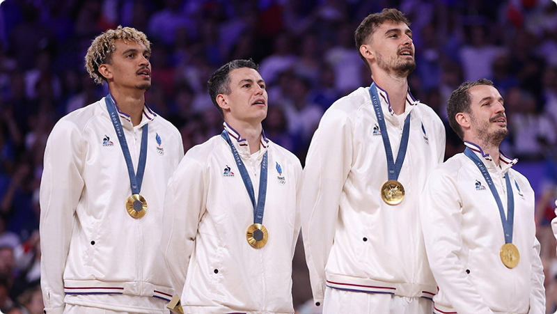 Four male athletes in white tracksuits stand side by side wearing gold medals. They appear to be on a podium with blurred spectators in the background.