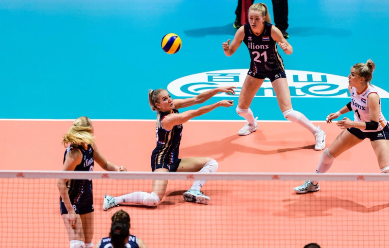 Volleyball player in a dark uniform dives to hit the ball while teammates watch during a match on an indoor court.