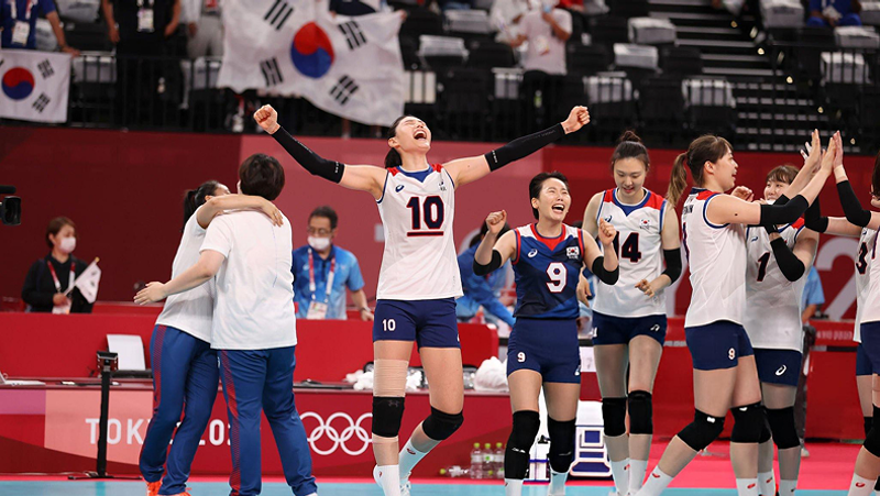 The image shows a women's volleyball team celebrating on the court, with the players wearing blue and white uniforms. The background is decorated with South Korean flags, and a "Tokyo 2020" banner is visible.