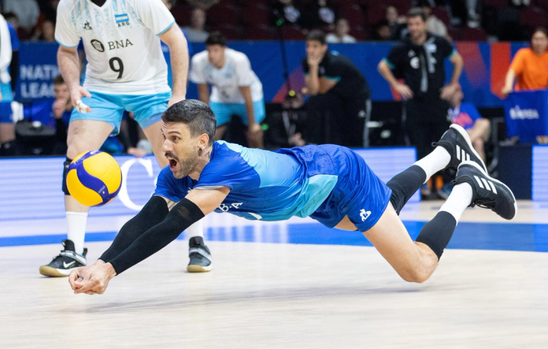 A volleyball player in a blue uniform dives to hit the ball during a match. Teammates and coaches are visible in the background.