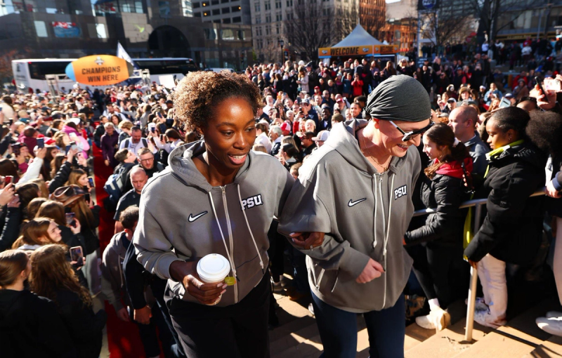 Two people in matching gray PSU jackets walking up steps through a large crowd. One is holding a coffee cup, and both are smiling. The crowd is cheering or taking pictures, with a "Champions Win Here" sign visible in the background.