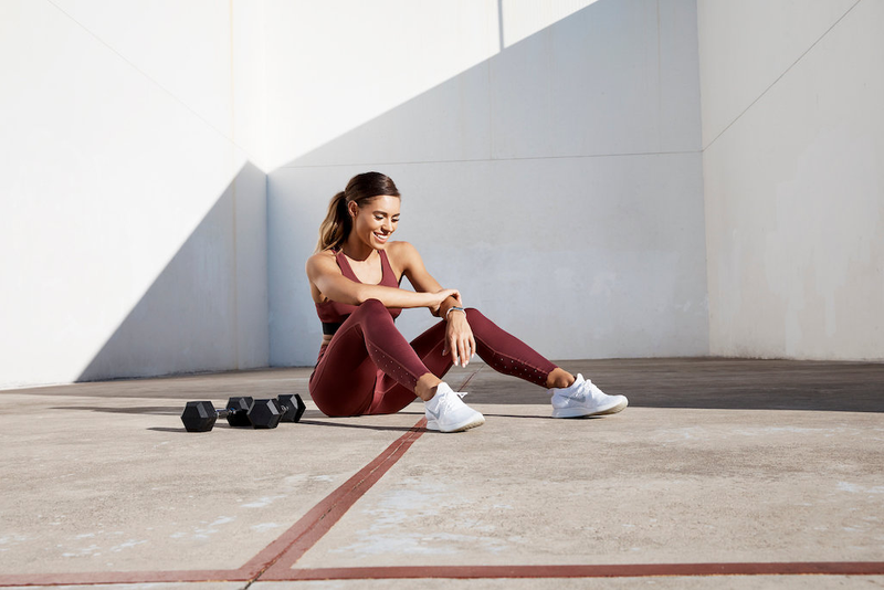 A woman in burgundy athletic wear sits on the ground in an outdoor gym setting, smiling and looking at her smartwatch. Two dumbbells are placed nearby.