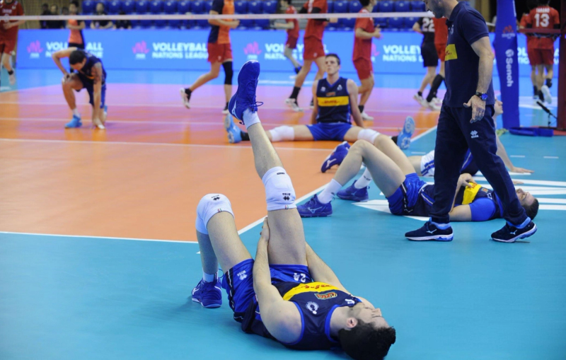 Volleyball players in blue uniforms are stretching on the court, preparing for a game. Some are lying on the floor holding their legs, while others are moving around. A coach or staff member in dark attire stands nearby.