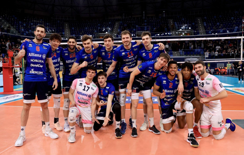 A volleyball team poses together on the indoor court. The players are wearing blue and pink uniforms with various logos, standing and kneeling in a group with smiles and gestures of camaraderie. The arena is filled with spectators in the background.