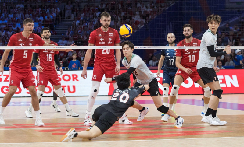 Volleyball players in action during a game, with one player in black diving to save the ball while others in red and white watch intently.