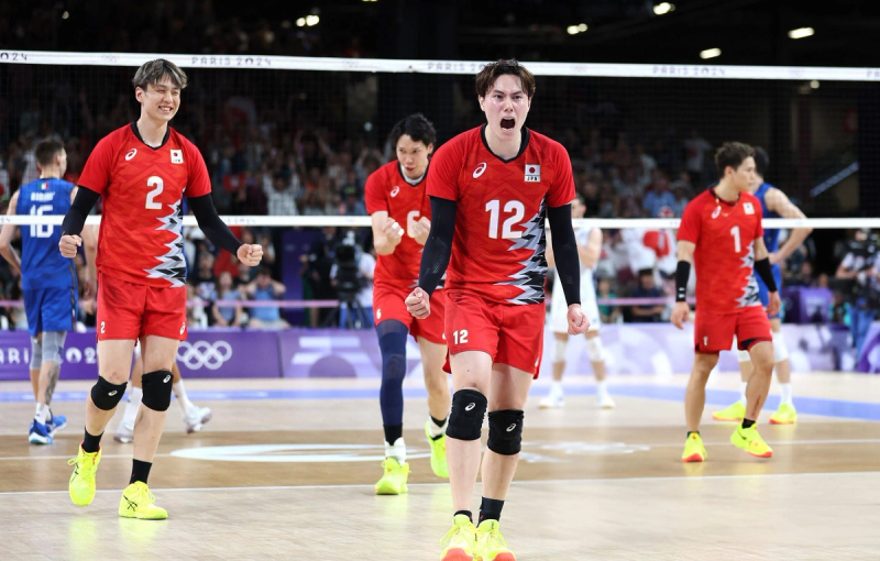 Men's volleyball players in red uniforms celebrate on an indoor court during a match, with a crowd in the background.