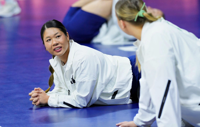 Two people in white jackets are lying on a sports floor, having a conversation. The background is out of focus.