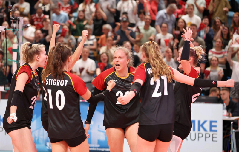 A group of female volleyball players huddles together in celebration on the court, with cheering fans visible in the background. They wear black and red uniforms, showing excitement and enthusiasm.