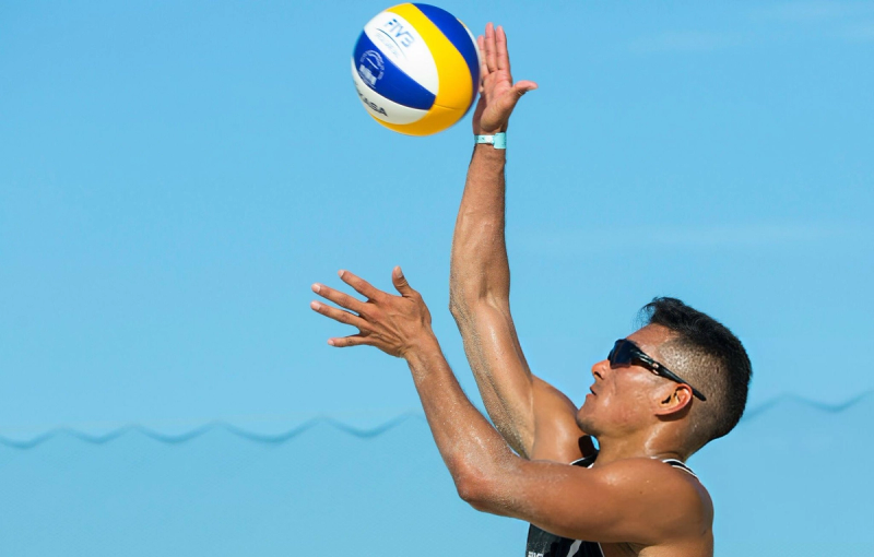 A beach volleyball player wearing sunglasses jumps to hit a volleyball against a clear blue sky.