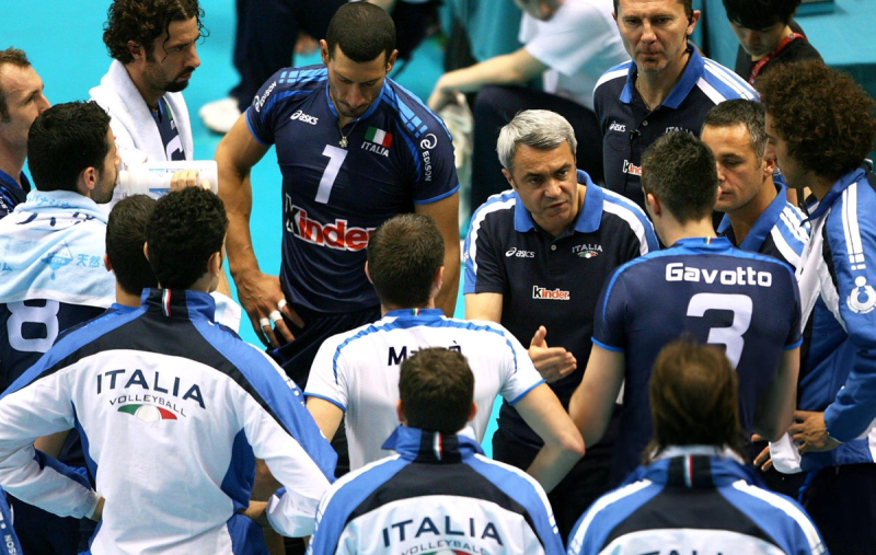 A volleyball coach is giving a strategy talk to his team, the Italian men's national volleyball team, during a game. The players are wearing blue and white jerseys, listening intently to the coach.