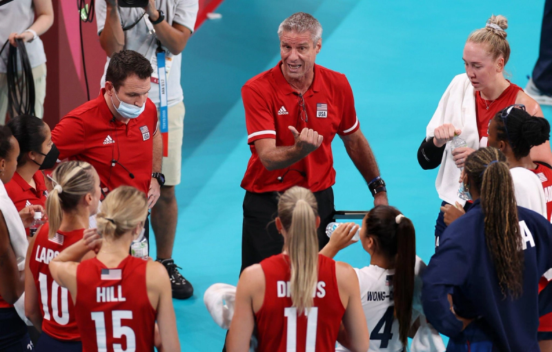 A coach gives a motivational talk to a women's volleyball team during a timeout. Players, wearing red jerseys, listen attentively while holding water bottles on the court.