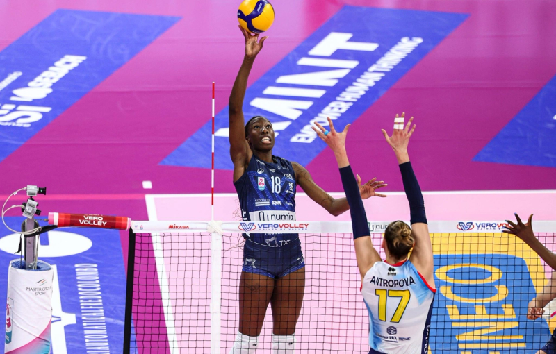 Volleyball player in a blue uniform jumps and reaches for the ball at the net, while an opponent in a white and blue uniform attempts to block. The court is pink and blue, with "VERO VOLLEY" branding visible.