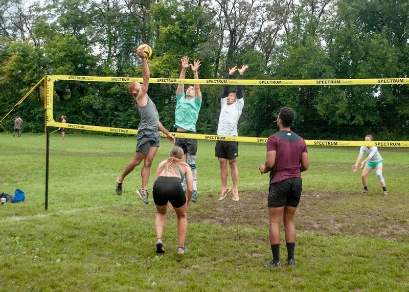 A group of people playing outdoor volleyball on a grassy field. One player jumps to hit the ball over the net, while others on the opposite side attempt to block. Spectators and teammates are in the background. Trees line the field.