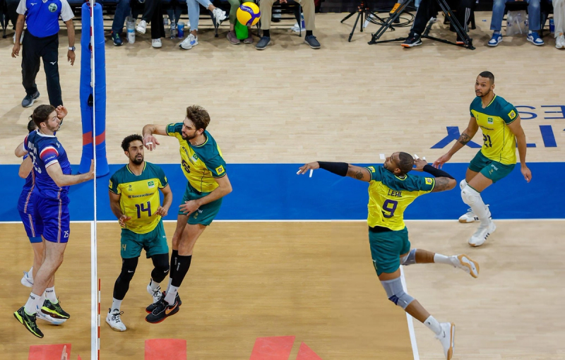 A volleyball match in action. A player in a yellow and green uniform is jumping to spike the ball, while teammates prepare nearby and opponents in blue are on the defensive side of the net.