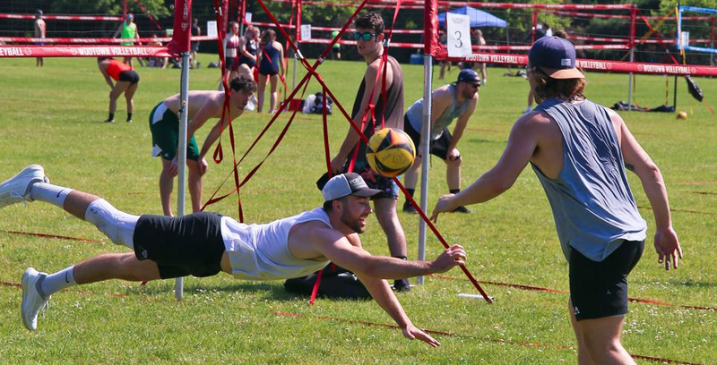 A group of people playing volleyball outside on a grass field. One player is diving to hit the ball, wearing a white tank top and cap. Other players watch and prepare for the next move. Volleyball nets and poles are visible in the background.