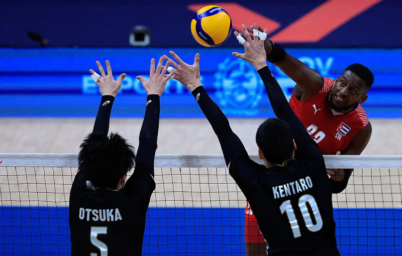 A volleyball player in a red jersey, number 23, spikes the ball over the net while two players wearing black jerseys attempt to block.