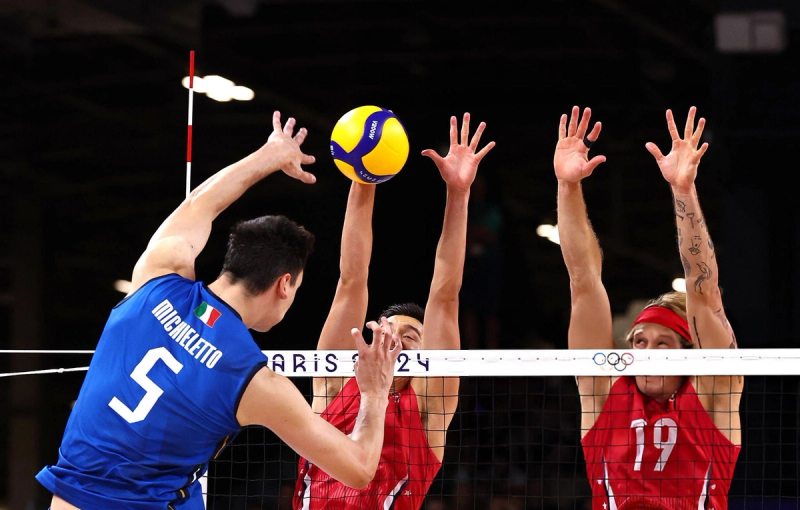 A volleyball player in a blue jersey prepares to spike the ball over the net against two opposing players in red jerseys who are jumping to block. The net is marked with "Paris 2024" and the Olympic rings logo.