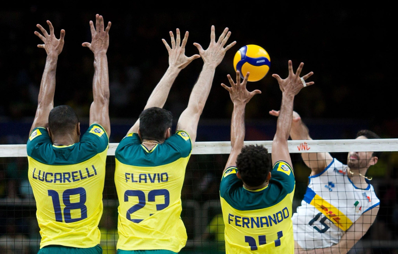 Three volleyball players in yellow jerseys jump to block a spike from a player in a white jersey on the opposite team at the net.