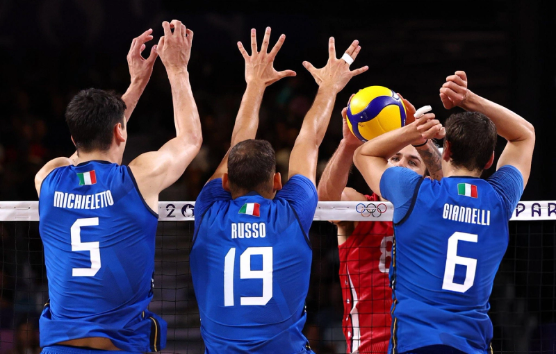 Volleyball players in blue jerseys block a player in a red jersey attempting to spike the ball at the net.