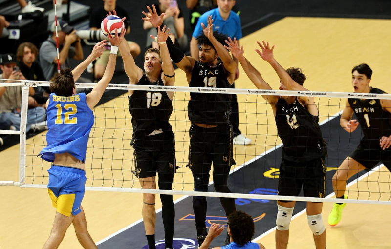 A volleyball game in action with a player from the Bruins team in a blue and yellow uniform attempting to hit the ball past three players in black jerseys from the opposing team. Spectators and a referee are visible in the background.