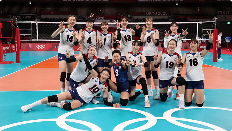 A volleyball team poses on the court at the Tokyo 2020 Olympics, smiling and flashing peace signs.