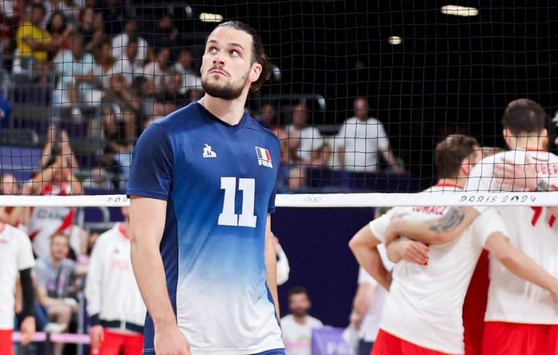 A volleyball player in a blue jersey with the number 11 stands on the court, looking up. In the background, a group of players in white jerseys huddle together near the net. The venue appears crowded with spectators.