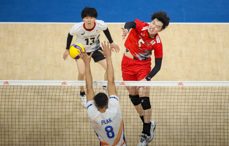 A volleyball player in a red uniform spikes the ball over the net, while an opponent in a white jersey attempts to block. Another player watches in the background.