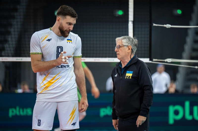 A volleyball player in a white and yellow uniform is talking to a coach wearing a black jacket with a small blue and yellow flag on it, in front of a volleyball net.