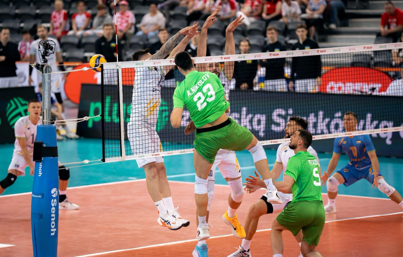 Volleyball players in action during a match, with one player in a green jersey jumping to spike the ball over the net as two opponents in white jerseys attempt to block. Spectators are visible in the background.
