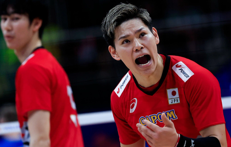 A volleyball player in a red jersey is shouting during a match. Another player is slightly blurred in the background. The jersey features the Japanese flag and various logos.