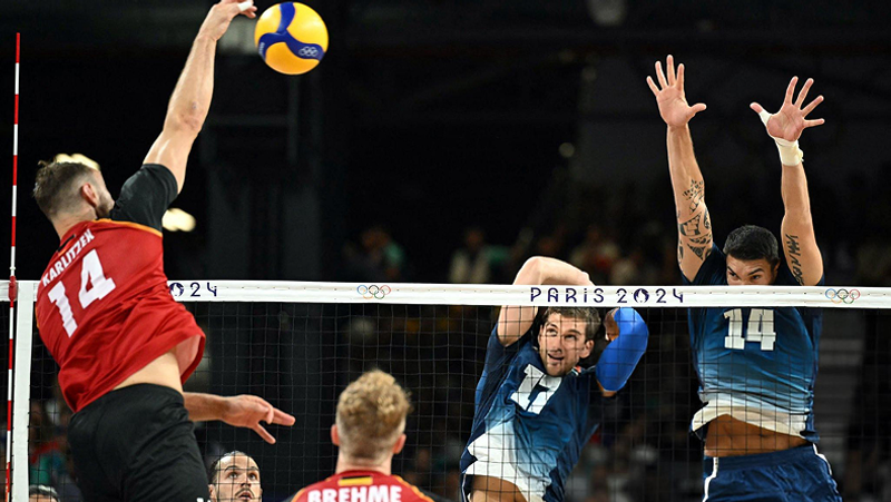 A volleyball player in red prepares to spike the ball over the net, while two players in blue jump to block during a Paris 2024 Olympic match.