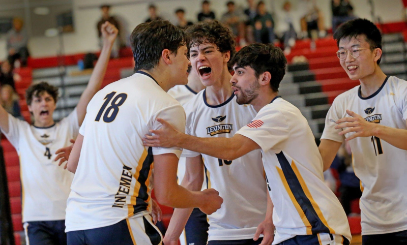 A group of male volleyball players celebrates enthusiastically on the court, wearing Lexington jerseys. They appear joyful and energetic, with one player's fist raised in the background.