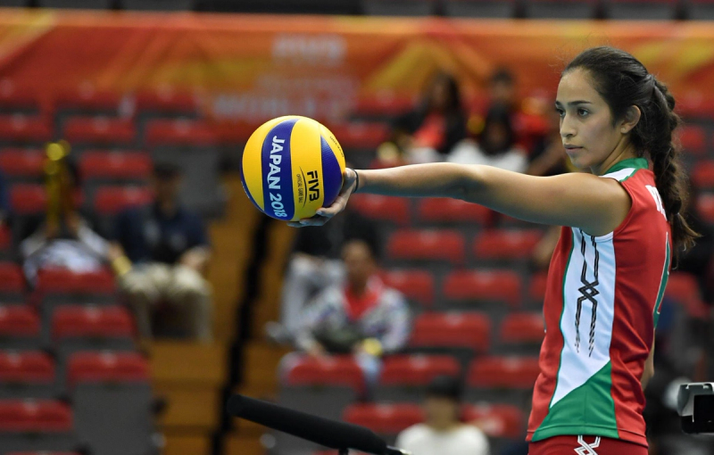 A volleyball player wearing a red, white, and green uniform prepares to serve, holding a blue and yellow FIVB 2018 ball. The background shows blurred spectators in a sports arena.