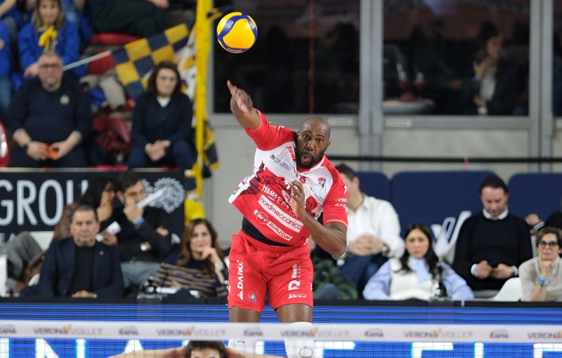 A volleyball player in a red uniform jumps to spike the ball during a match, with spectators watching from the stands in the background.