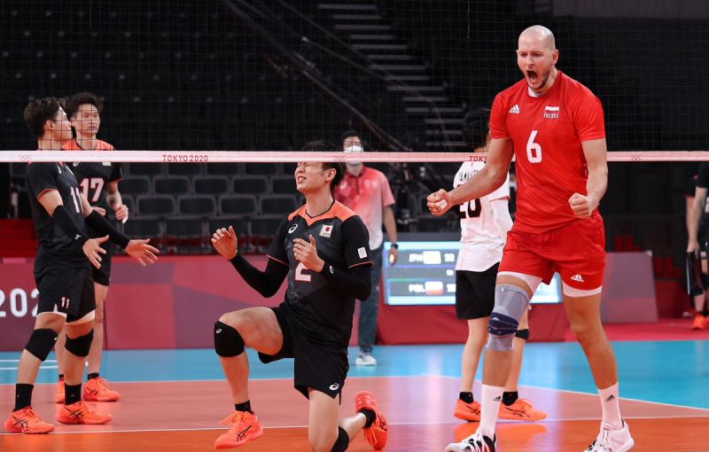 Volleyball players in action during a match at the Tokyo 2020 Olympics. A player in red celebrates with clenched fists while a player in black kneels with a pained expression. The scoreboard in the background shows scores for Japan and Poland.