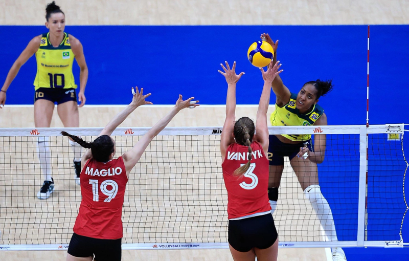 A volleyball match in action, with a player in a yellow jersey spiking the ball over the net. Two players in red jerseys attempt to block. Another player in a yellow jersey stands ready in the background.