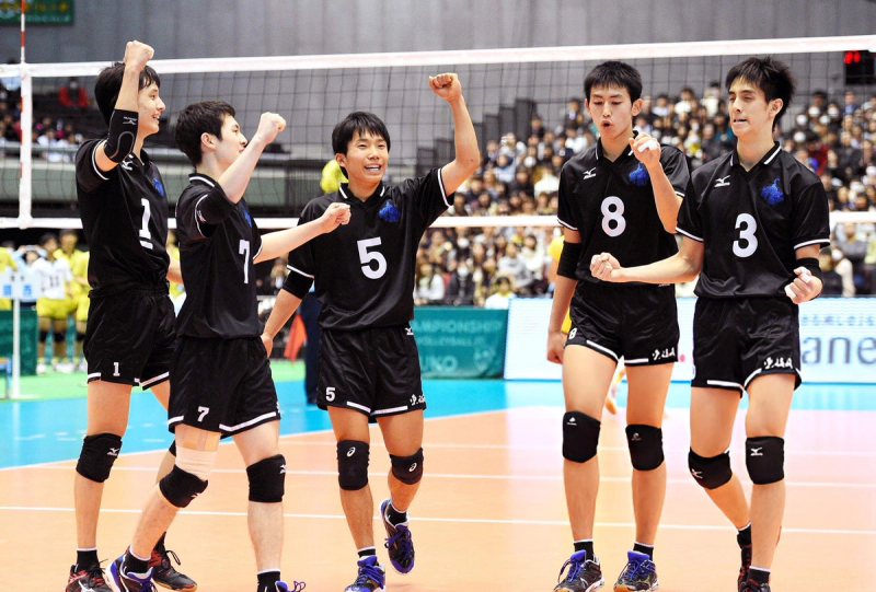 Five volleyball players in black uniforms celebrating on the court with fists raised, showing enthusiasm and unity.