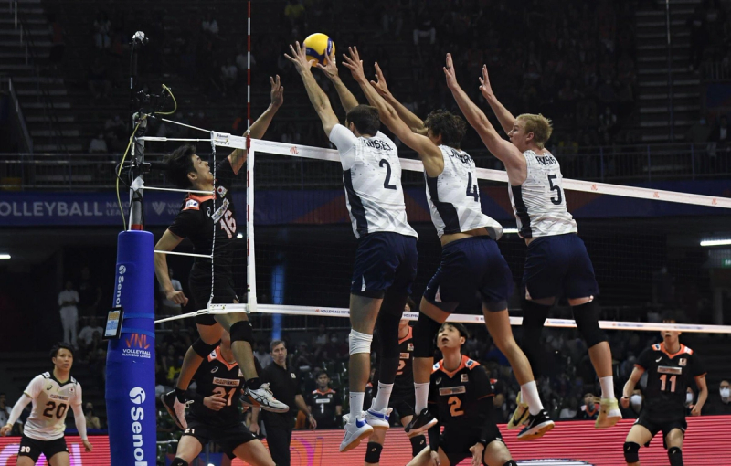 Volleyball players in action; a player in a black jersey jumps to spike the ball as three players in white jerseys attempt to block it at the net during a match.