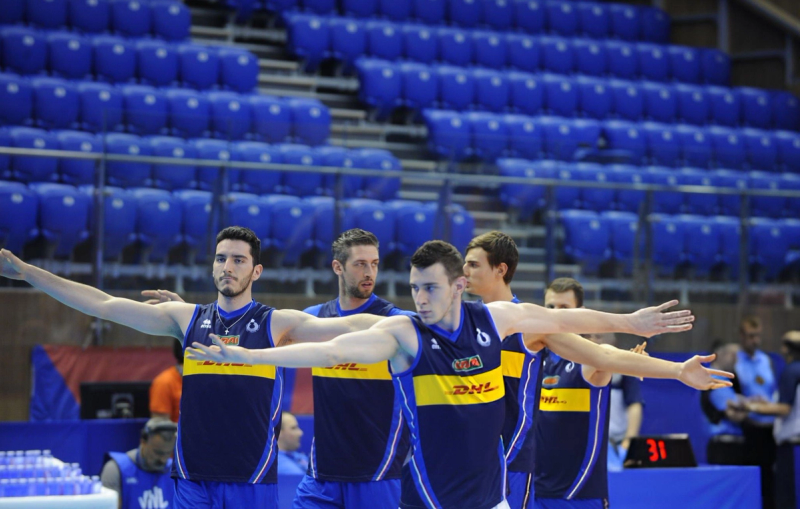 Volleyball players in blue and yellow uniforms stand in a row with arms extended in a stadium.