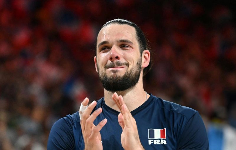 A man with a beard and tied-back hair claps while appearing emotional. He is wearing a dark blue shirt with a French flag and "FRA" on it. The background is blurred with a jumble of red and other colors.
