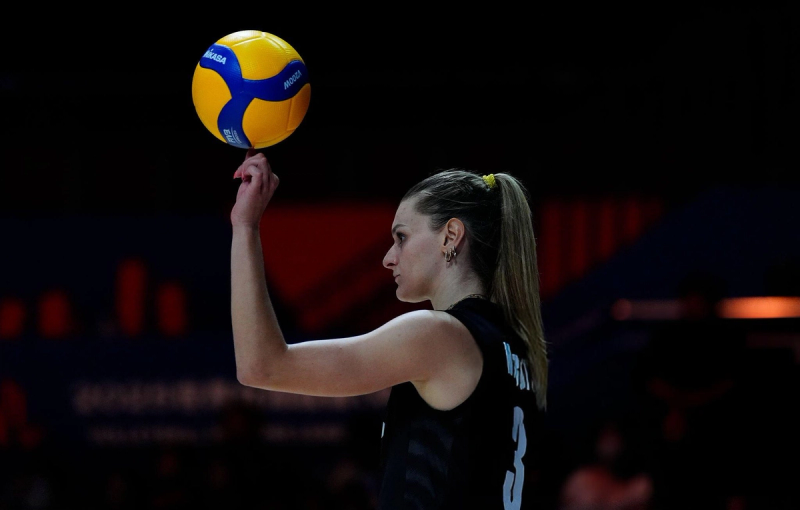 A volleyball player balances a ball on her fingertip during a game, wearing a black jersey with the number 3.